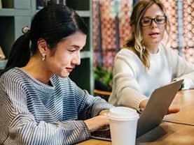 Korean woman working on laptop and having coffee, sitting next to a colleague.